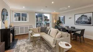 A modern living room with a sectional sofa, a dining table, and chairs. There is a large window providing a view of the outside. - JOIVY Astonishing flats near Mayfair & Piccadilly Circus (London)