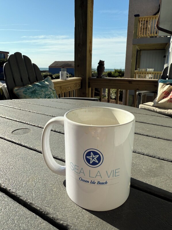 Caneca branca com a inscrição 'SEA LA VIE Ocean Isle Beach' sobre um deck de madeira.