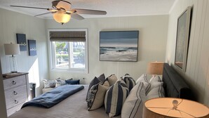 Une chambre à coucher avec un lit, des oreillers, un ventilateur de plafond, une fenêtre avec des stores et un tableau au mur.