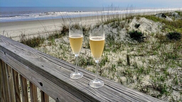 Deux coupes de champagne posées sur une rambarde en bois, donnant sur une plage avec des dunes de sable et l’océan en arrière-plan.