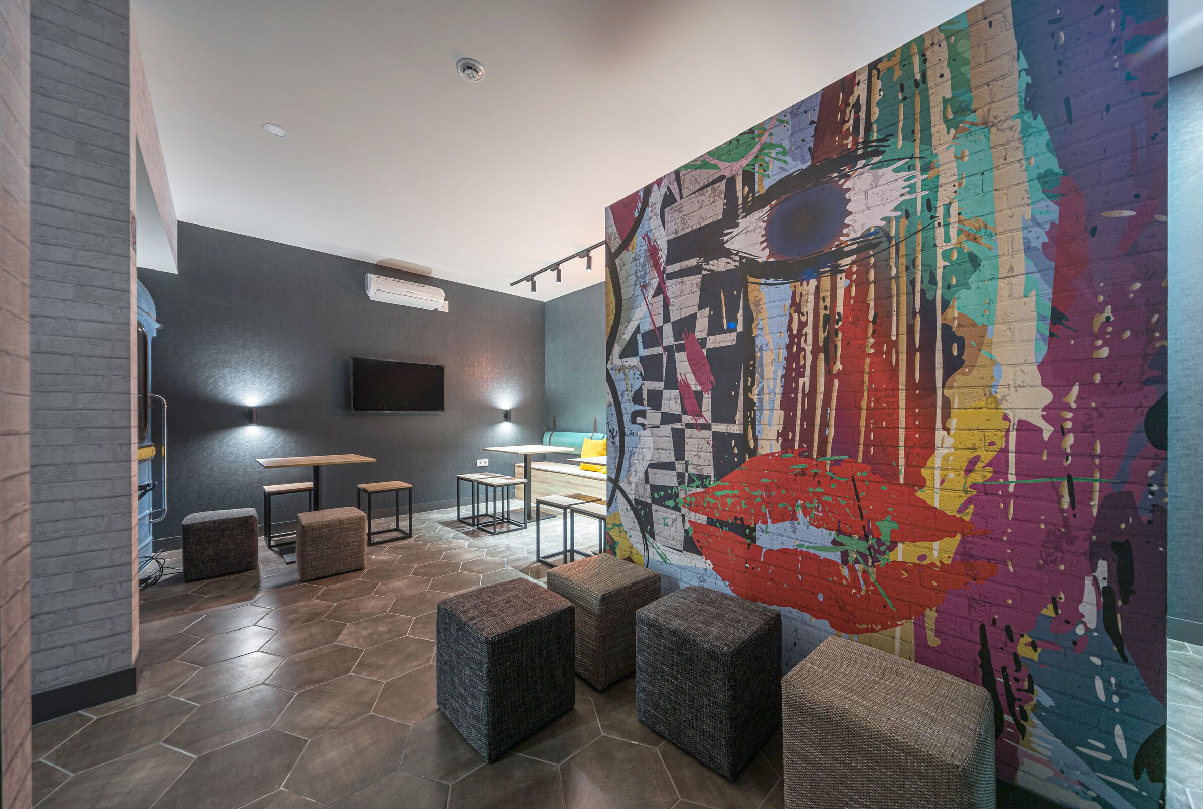 Un espace de détente moderne doté d’un sol à motifs hexagonaux, de plusieurs options d’assises, d’une grande fresque murale abstraite et d’un téléviseur à écran plat fixé au mur.