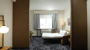 A hotel room with a bed, a desk, a chair, a lamp, a window with curtains, and a wall-mounted air conditioner.