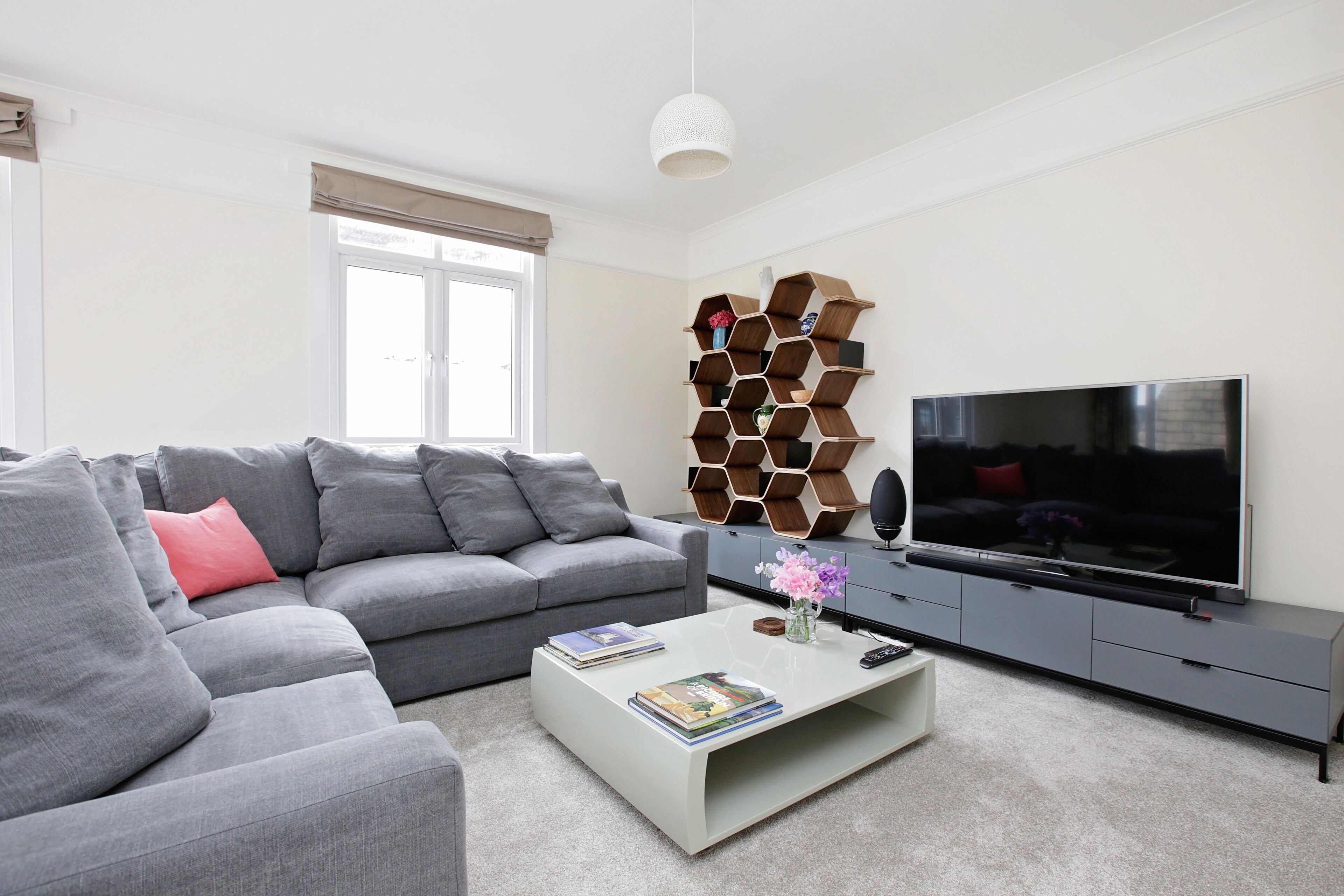 Una sala de estar moderna con un sofá gris, una mesa de centro con libros y un florero, un televisor sobre un mueble gris, y una decoración de pared de madera original.