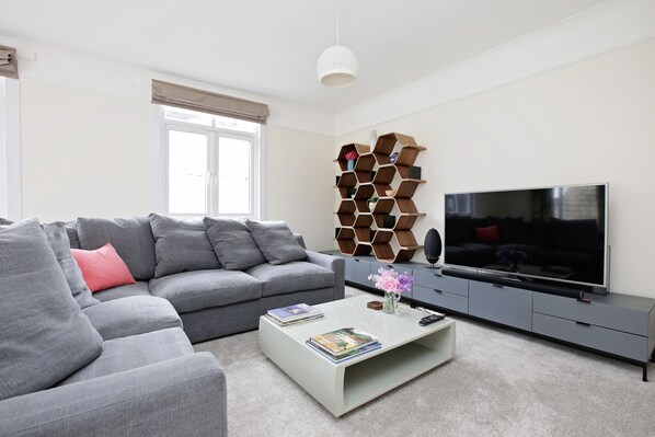 A modern living room with a grey sofa, a coffee table with books and a vase, a TV on a grey cabinet, and a unique wooden wall decor.