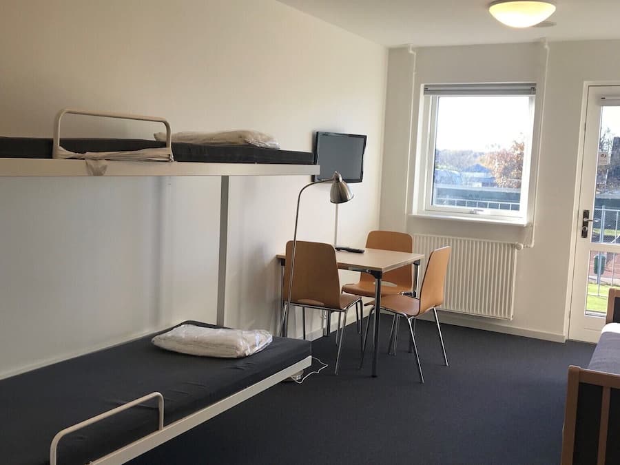 A dormitory room with bunk beds, a desk, chairs, a computer monitor, and a window with a view.
