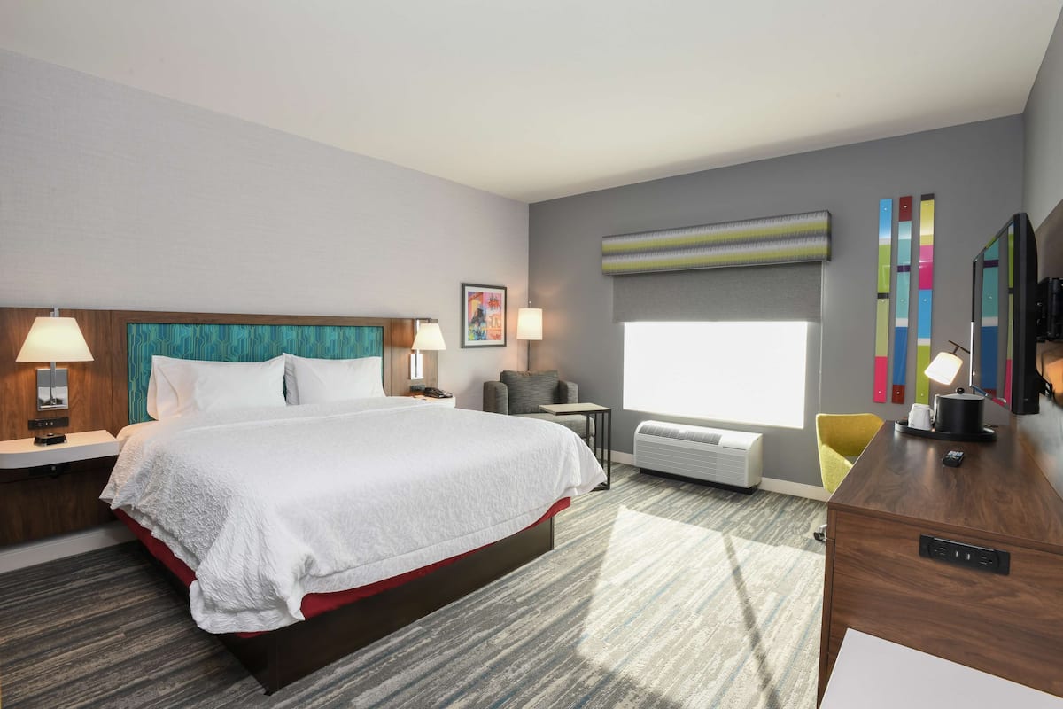 A hotel room with a large bed, bedside tables with lamps, a desk, a chair, a large window, and a wall-mounted air conditioner.