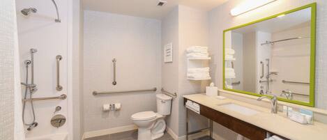 A modern bathroom with a shower, toilet, and sink, equipped with grab bars and a large mirror.