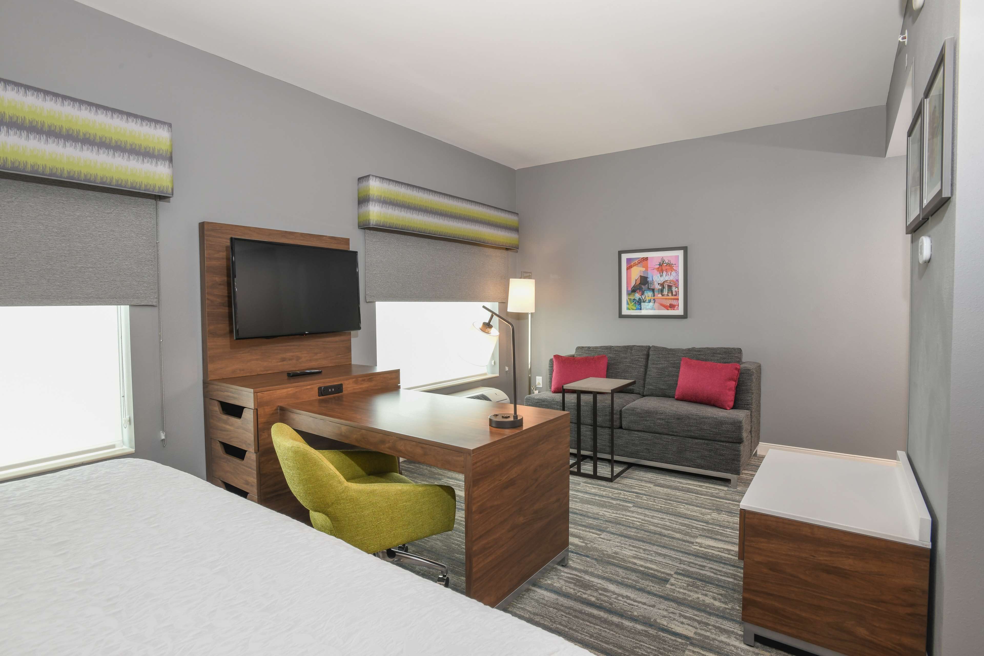 A hotel room with a bed, a desk with a chair, a sofa, a TV, and a lamp.