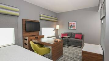A hotel room with a bed, a desk with a chair, a sofa, a TV, and a lamp.