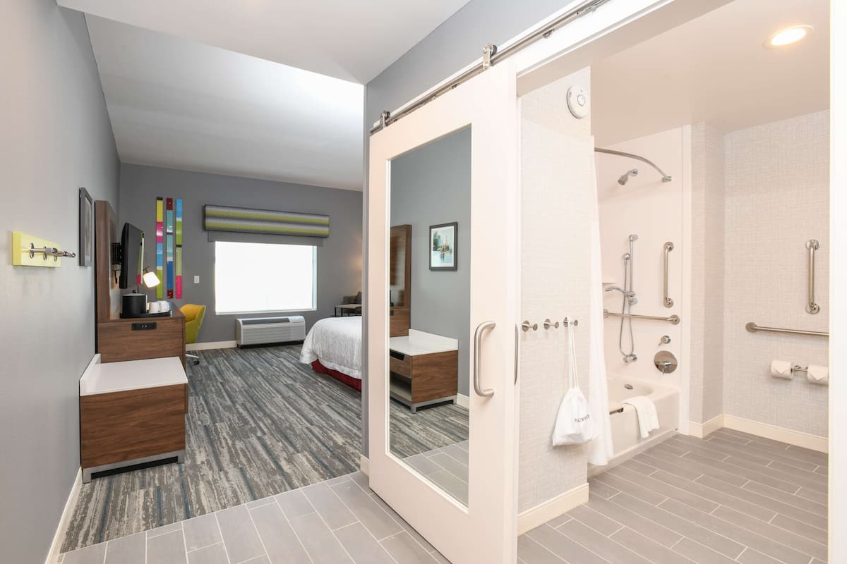 A modern hotel room with a large bed, a walk-in closet, a bathroom with a shower, and a view of the city.