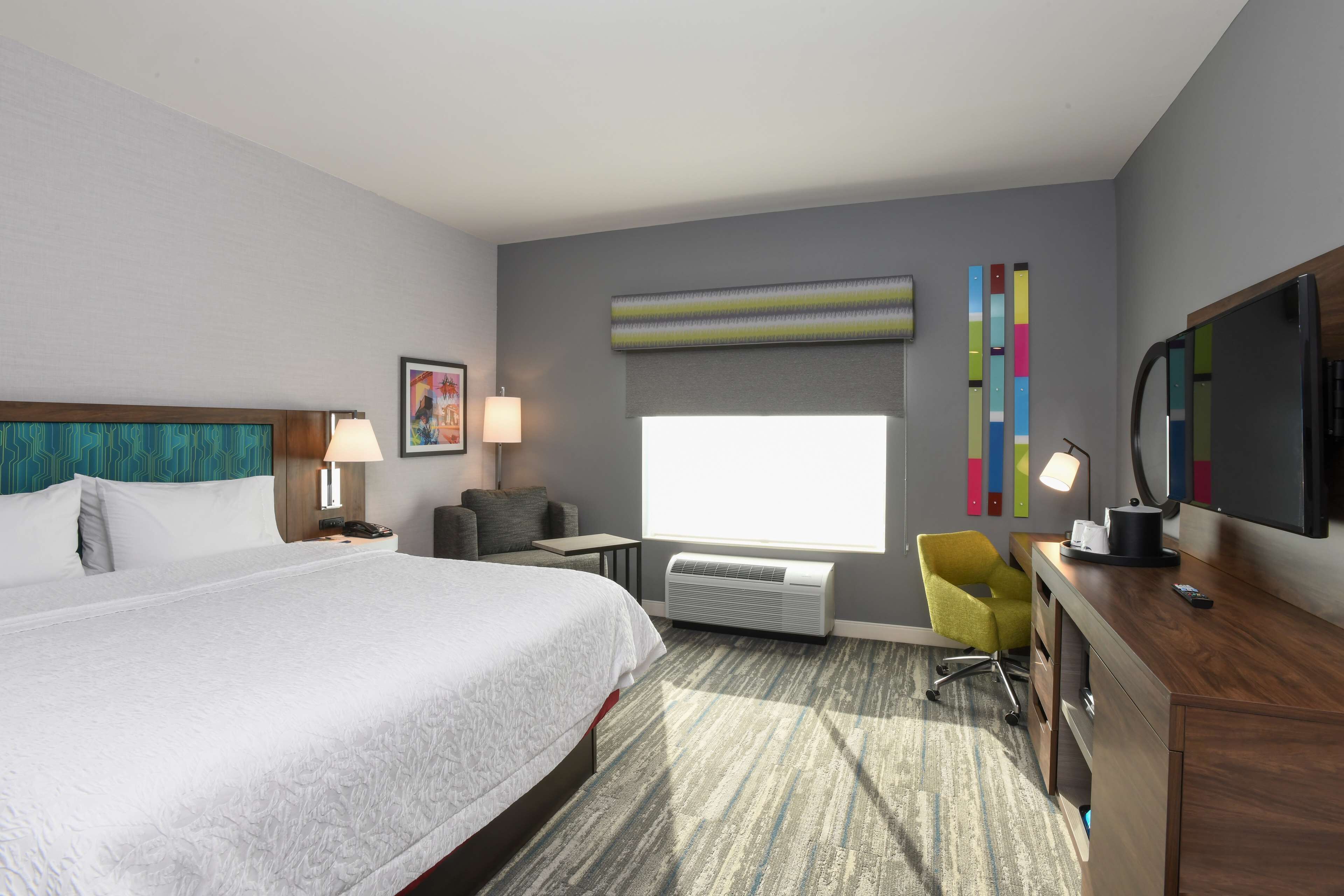 Habitación de hotel con una cama grande, un escritorio, una silla, un televisor y una ventana con persianas.
