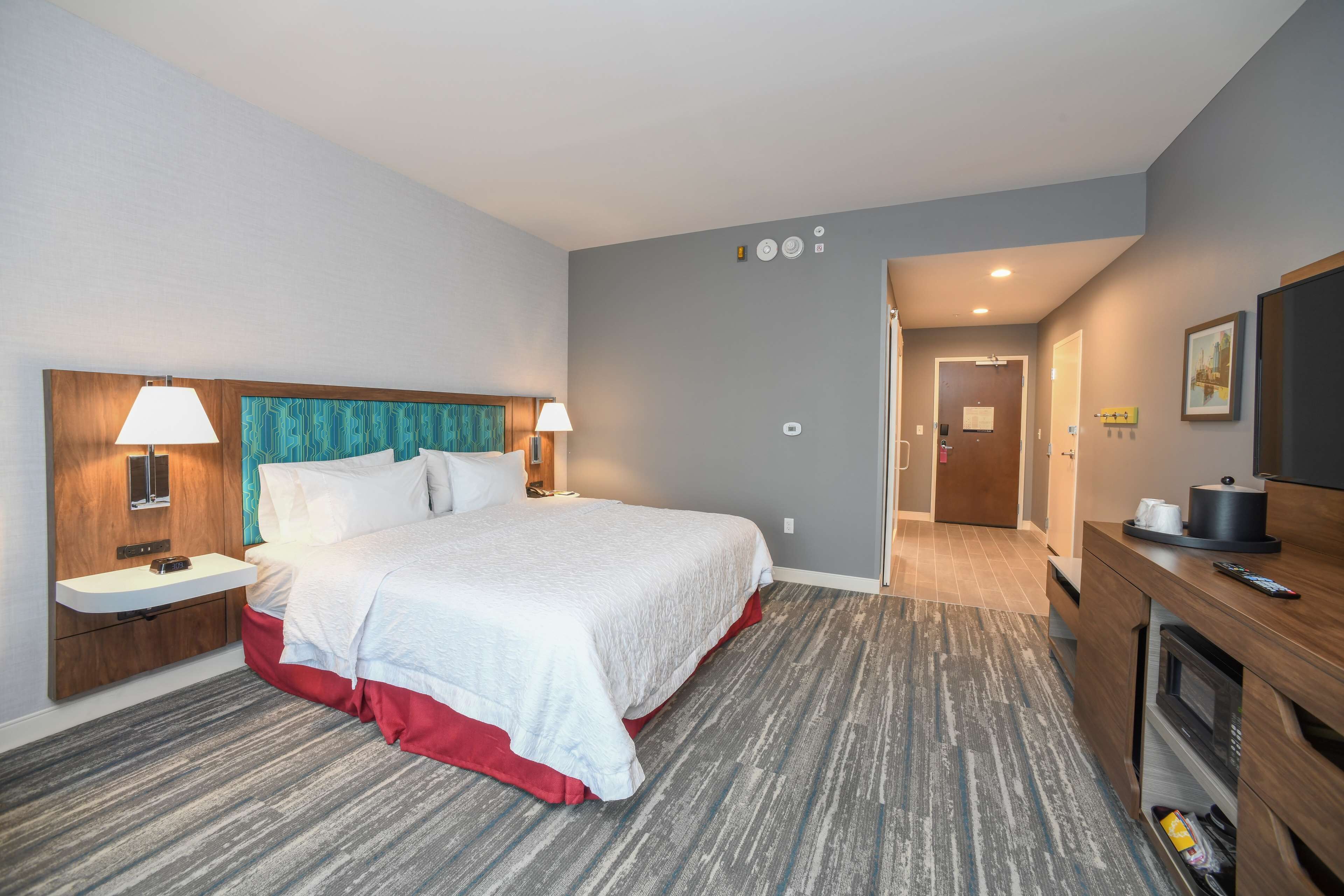 Une chambre d’hôtel avec un grand lit, des tables de chevet avec des lampes, une tête de lit en bois, un téléviseur intégré, un four à micro-ondes et une petite kitchenette.