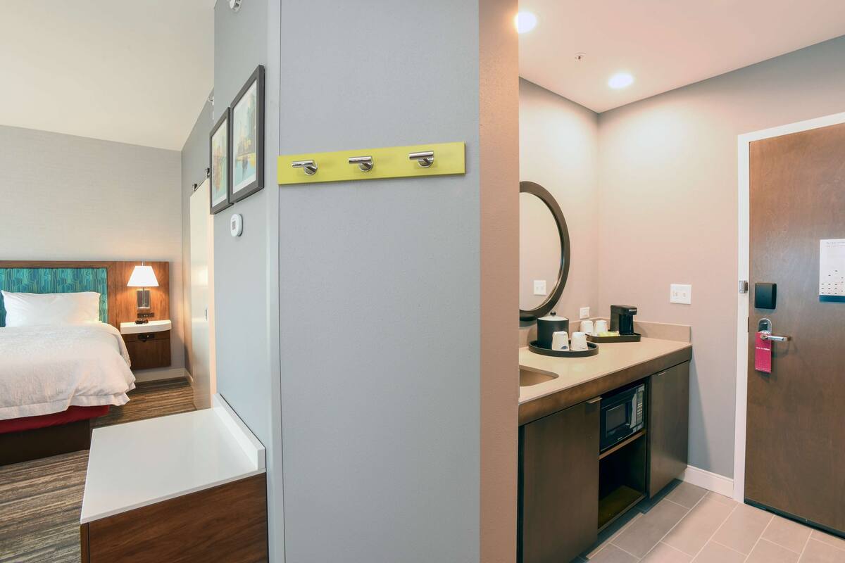 A hotel room with a bed, bedside table, lamp, and a small kitchenette with a microwave.