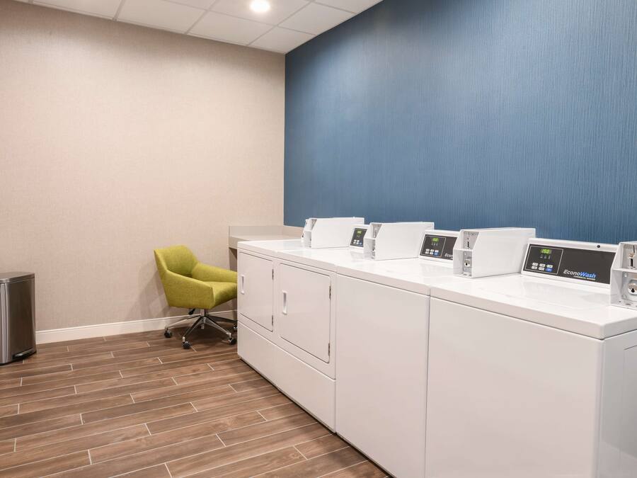 A laundry area with a row of washing machines, a trash can, and a green chair.