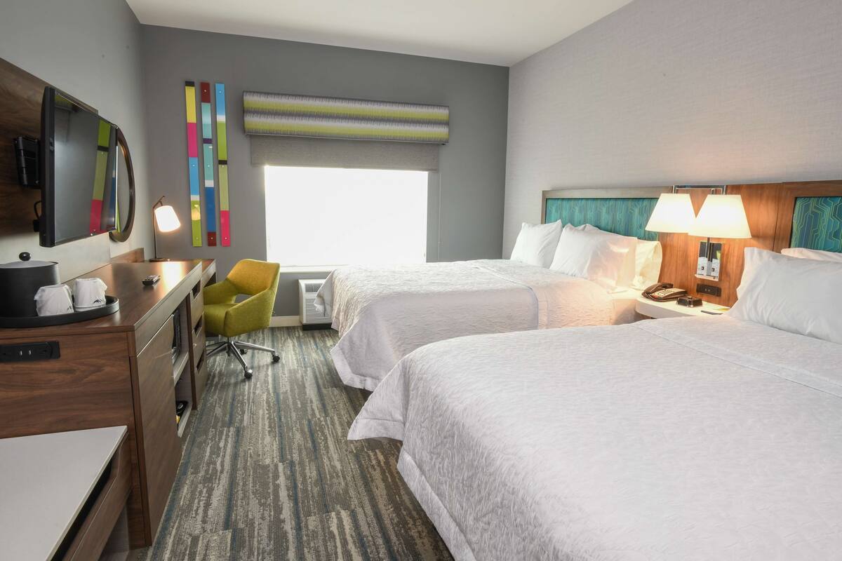 A hotel room with two beds, a desk, a chair, a TV, and a window with blinds.