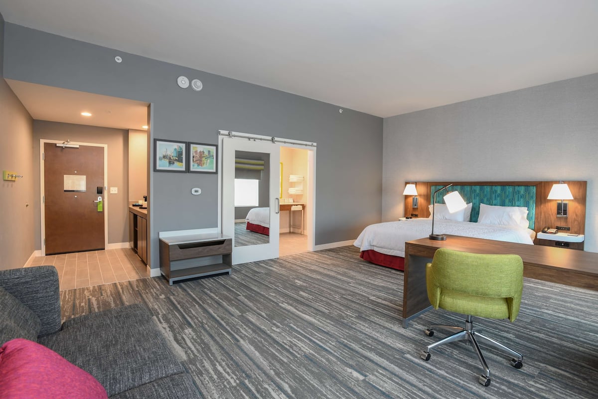 A hotel room with a bed, a desk, a chair, a bathroom, and a hallway leading to another room.
