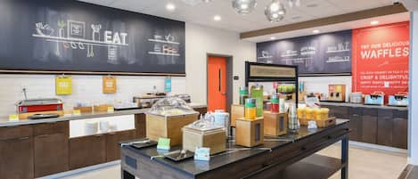 A breakfast buffet area with various food stations, including a waffle station, and a chalkboard menu.