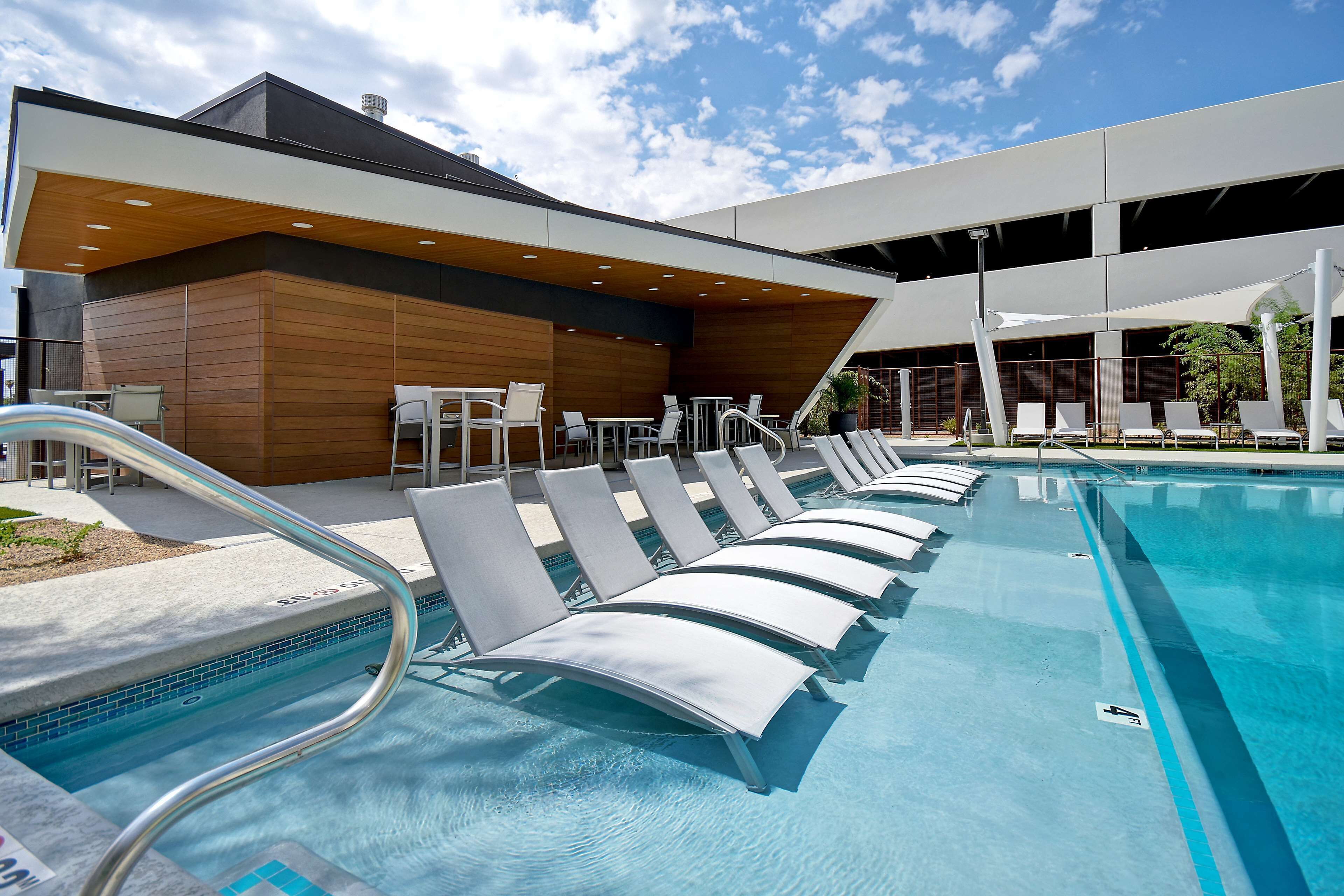 Un espace piscine moderne, doté de chaises longues et de tables, une piscine à l’eau d’un bleu limpide, et un bâtiment à la façade en bois et aux avant-toits saillants.