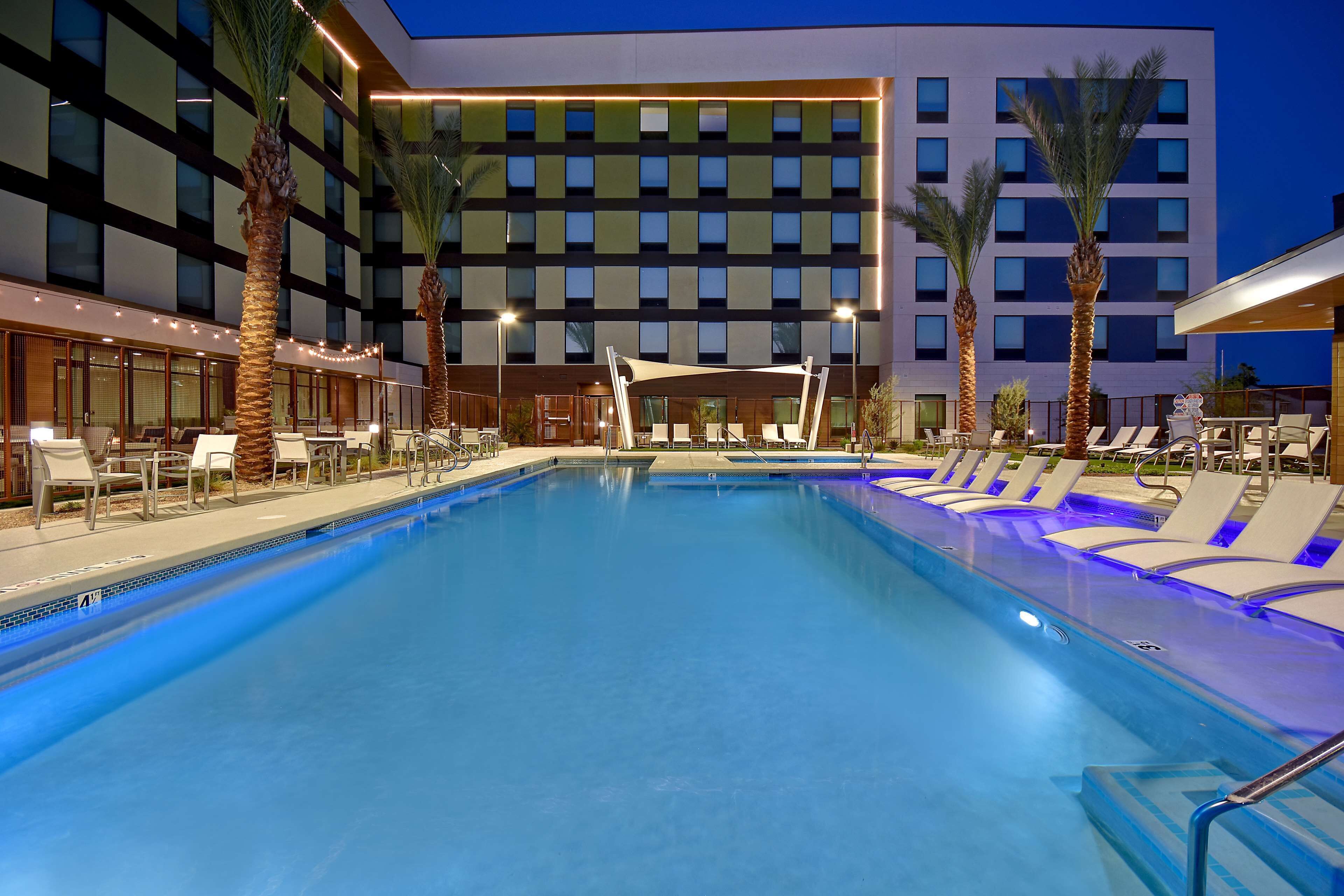 A swimming pool with lounge chairs and palm trees, surrounded by a modern building with multiple floors and balconies.