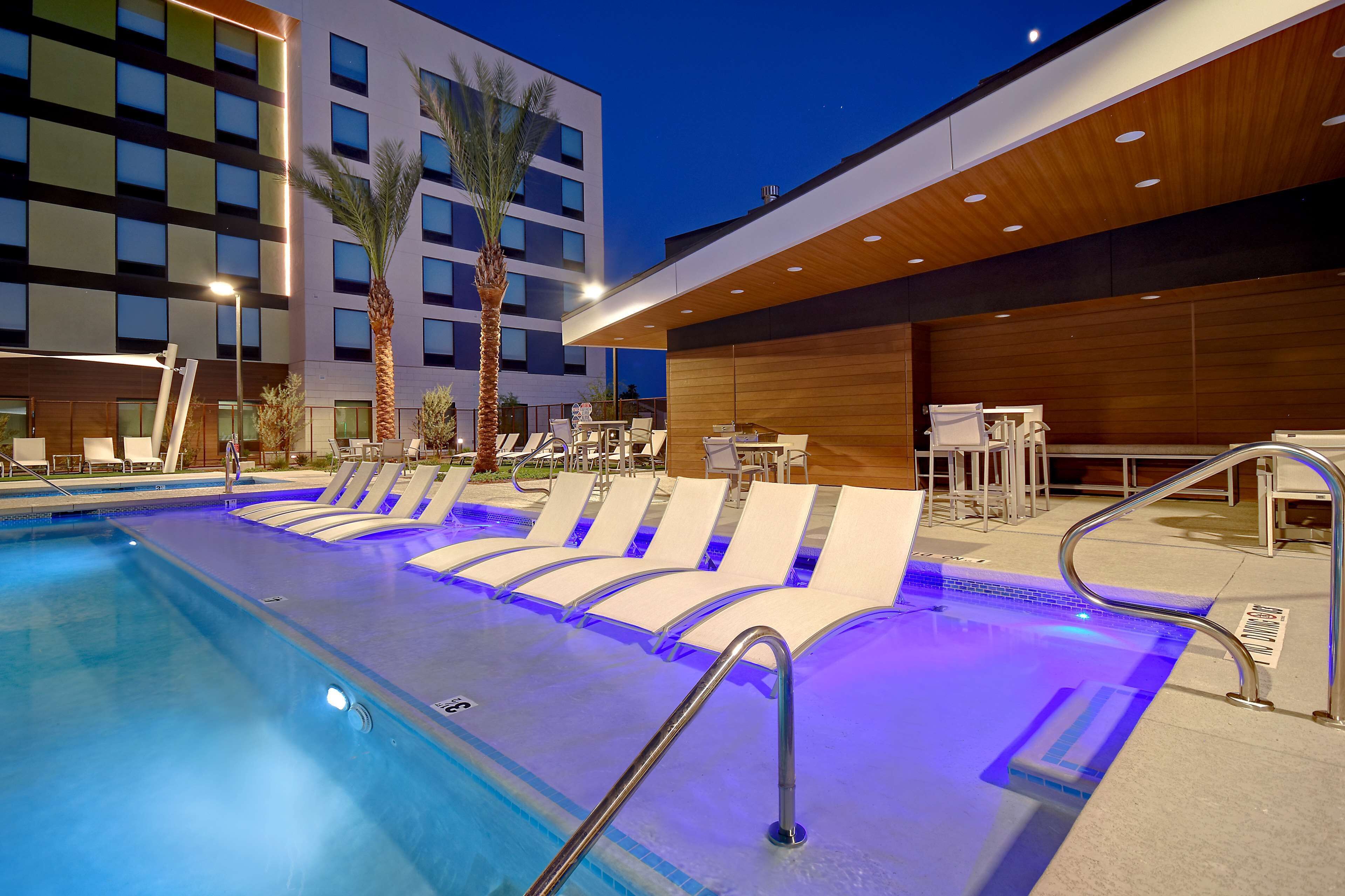 A modern hotel pool area with lounge chairs, a palm tree, and a building in the background.