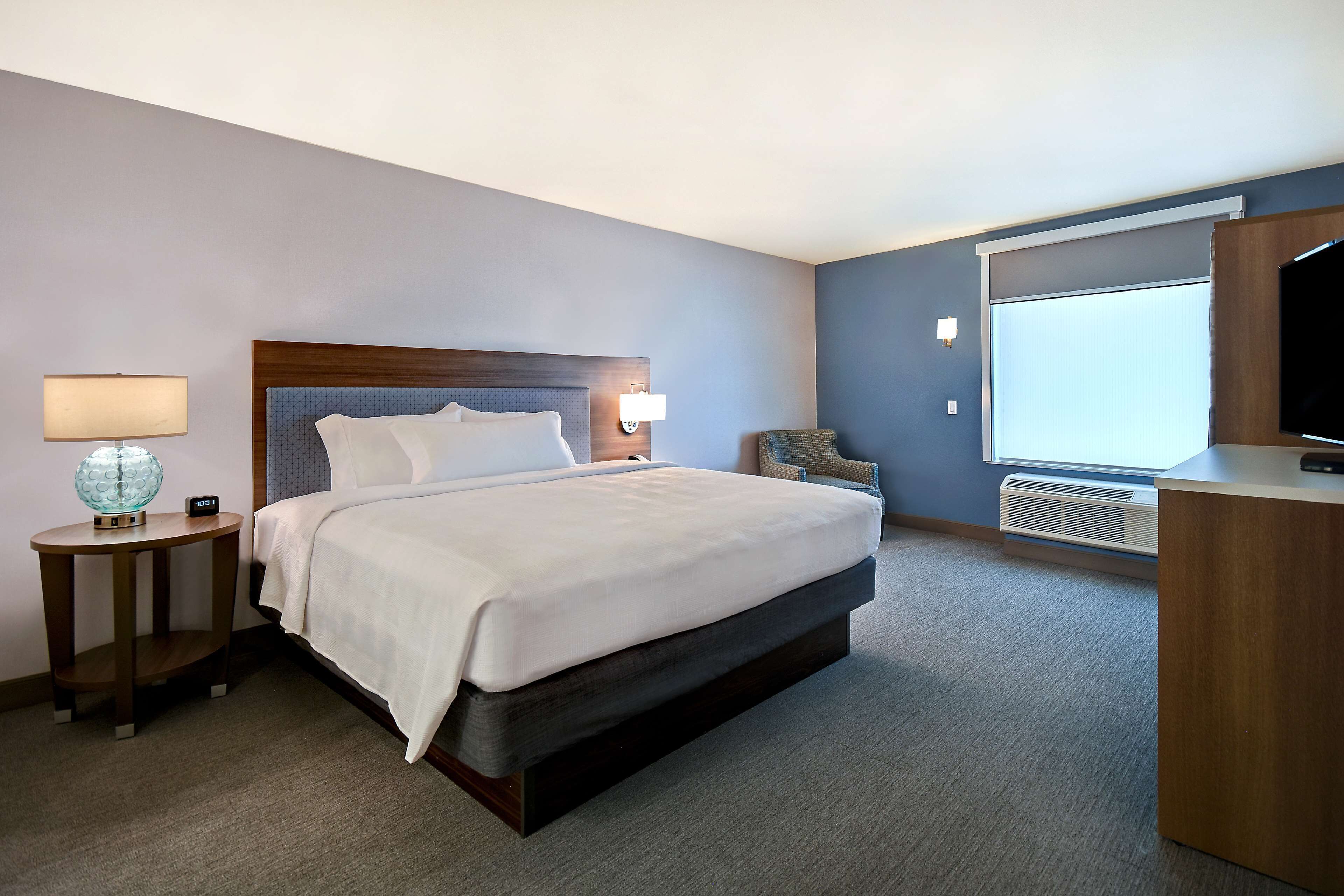 Une chambre d’hôtel comprenant un lit, une table de chevet avec une lampe, une chaise, un bureau avec une télévision et une grande fenêtre.