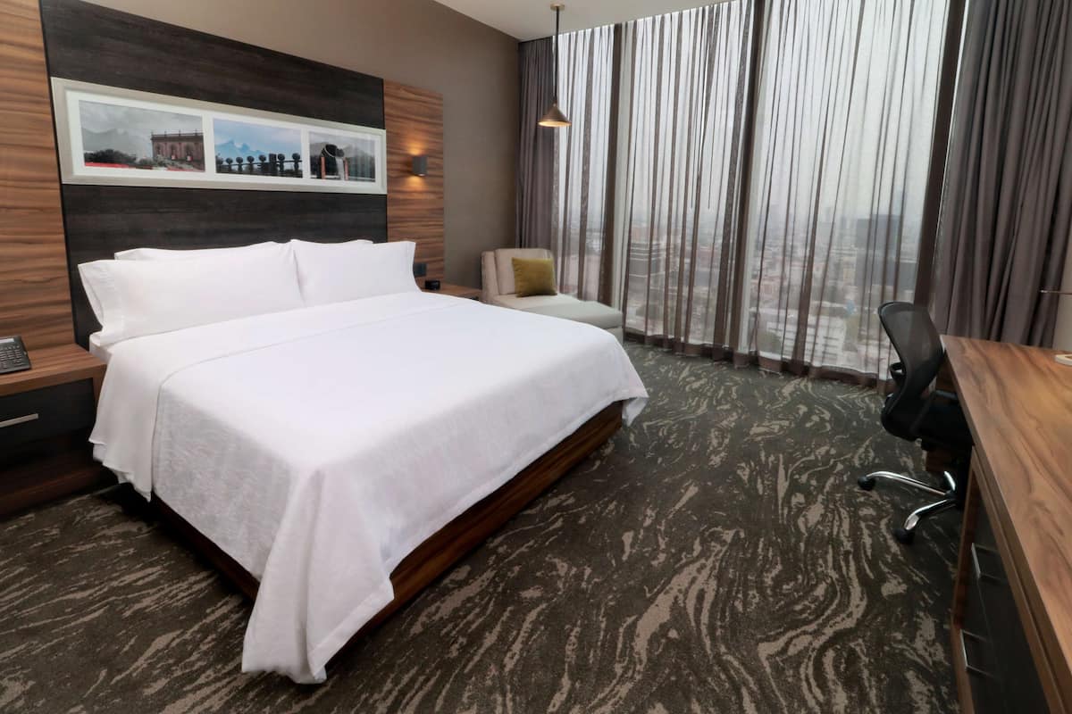 A hotel room with a large bed, a desk, and a chair. There is a window with curtains and a view of buildings.