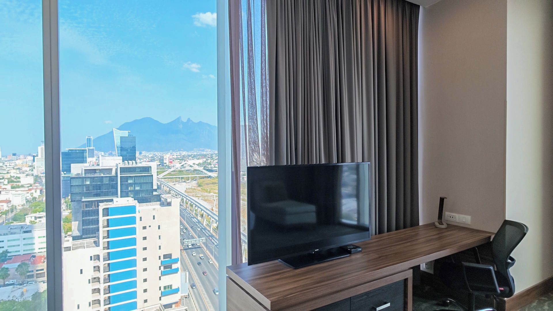 A hotel room with a large window, a flat-screen TV, a wooden desk, and a chair.