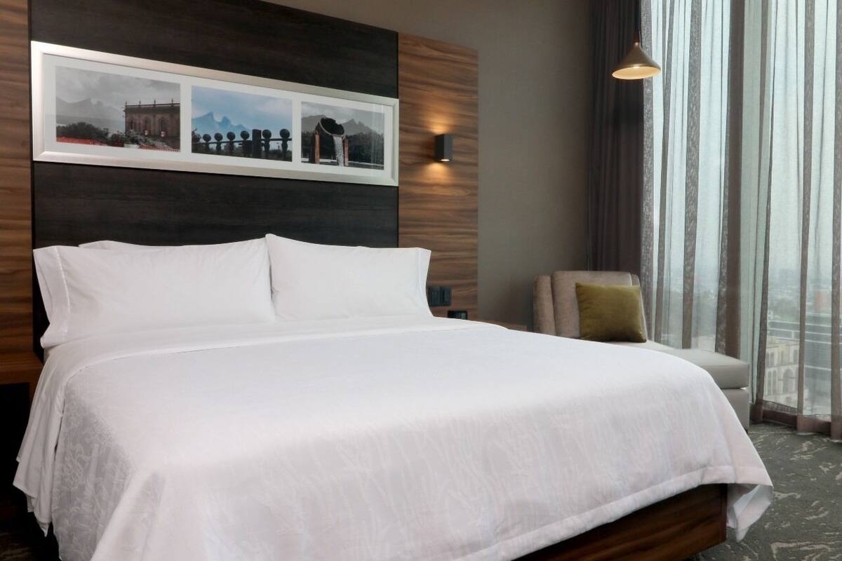 A hotel room with a large bed, a headboard with a picture, a side table, and a window with curtains.