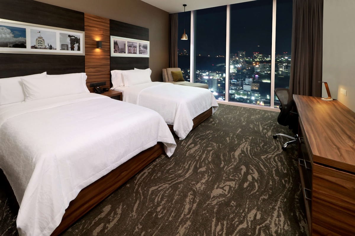 A hotel room with two beds, a desk, and a large window offering a city view at night.