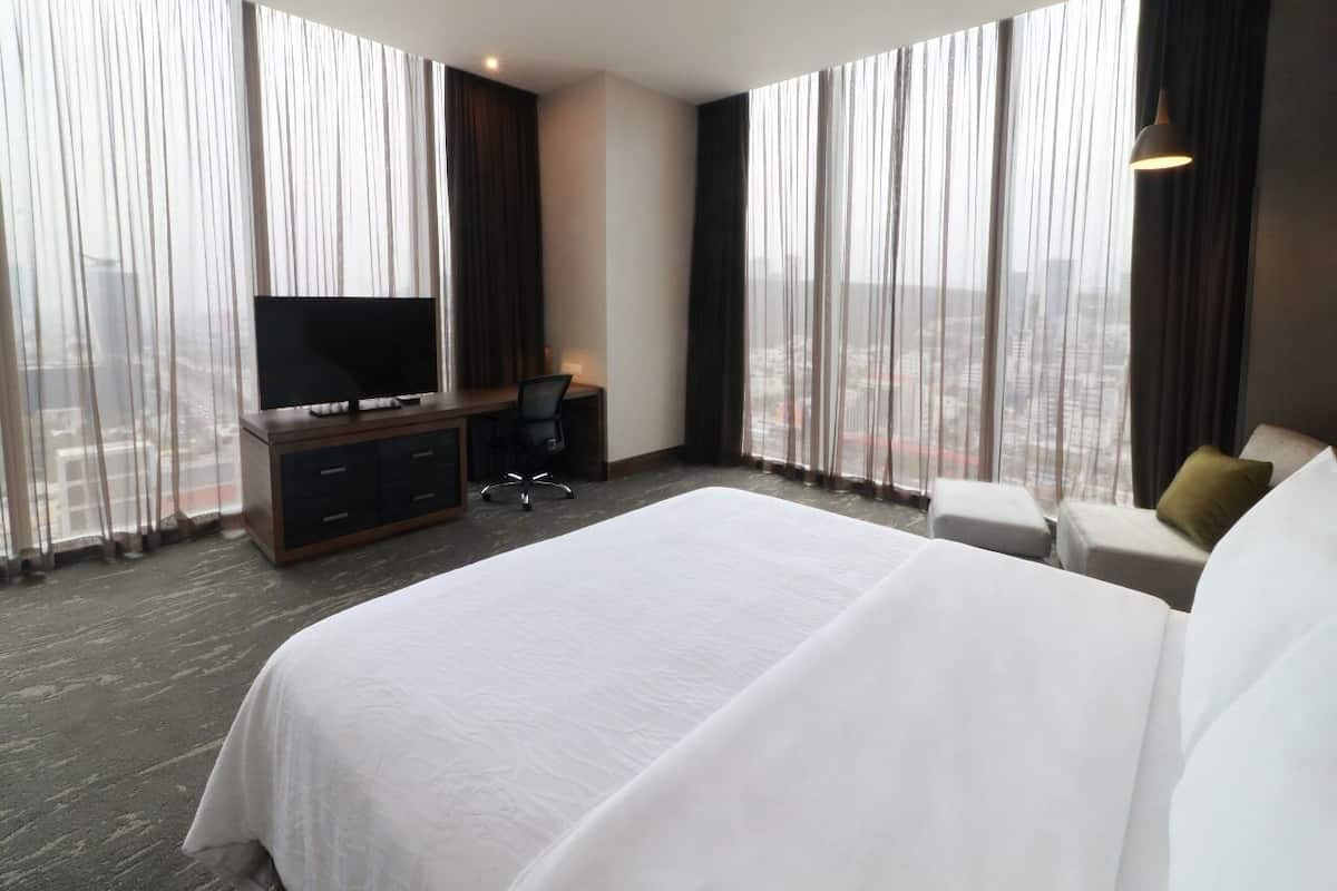 A hotel room with a large bed, a desk with a TV, and a view of the city through floor-to-ceiling windows.