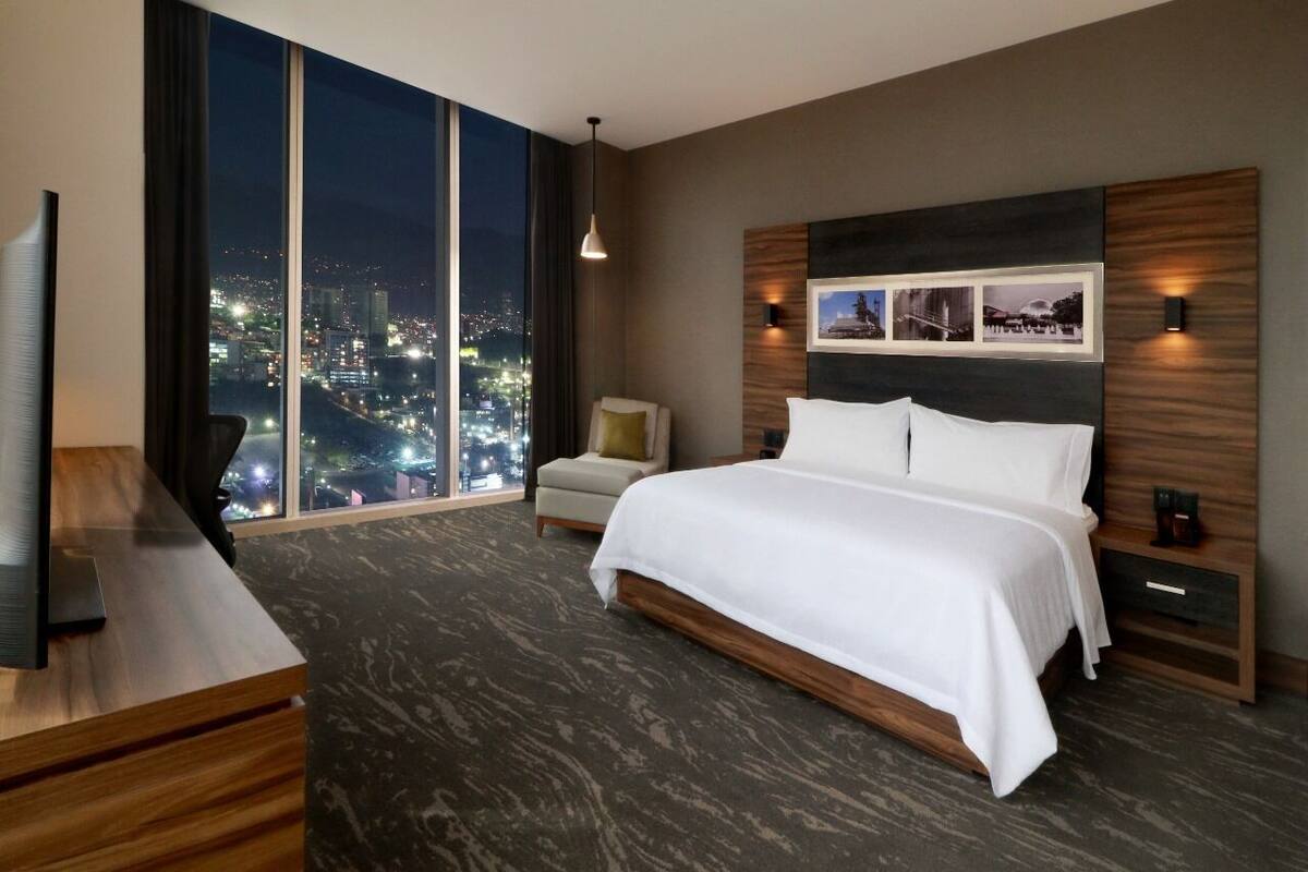A modern hotel room with a large bed, a desk, a chair, and a city view.