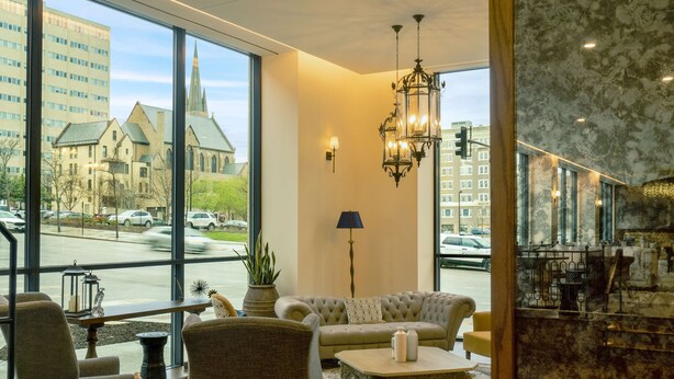A modern hotel lobby with large windows, a chandelier, and a seating area with a sofa and armchairs.