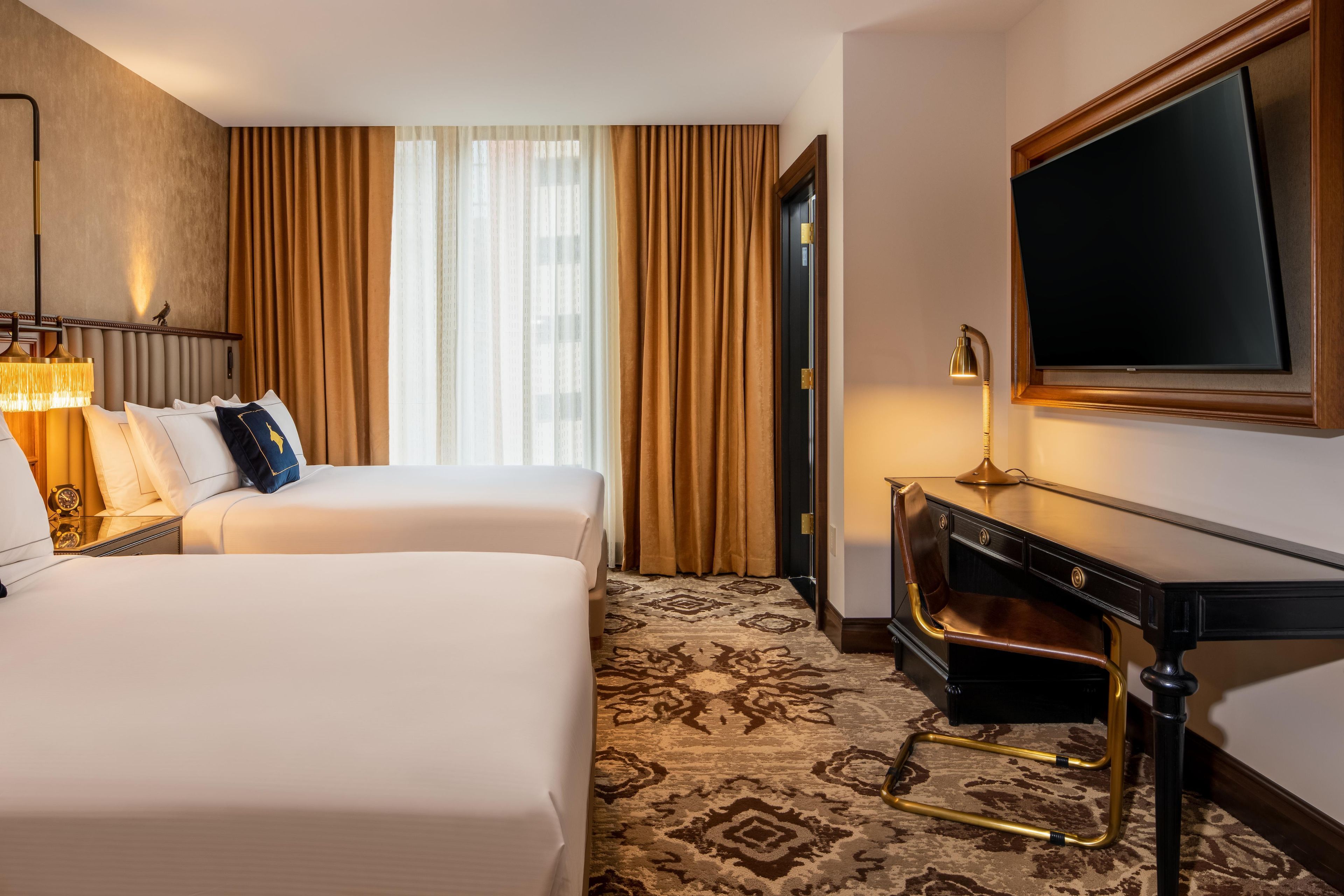 A hotel room with two beds, a desk, a television, and a large window with curtains.