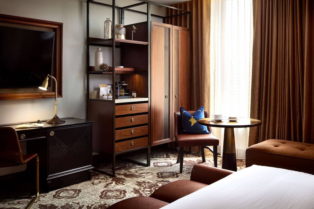 A hotel room with a bed, a desk with a lamp, a chair, a small table with a cup and saucer, and a wardrobe with shelves.
