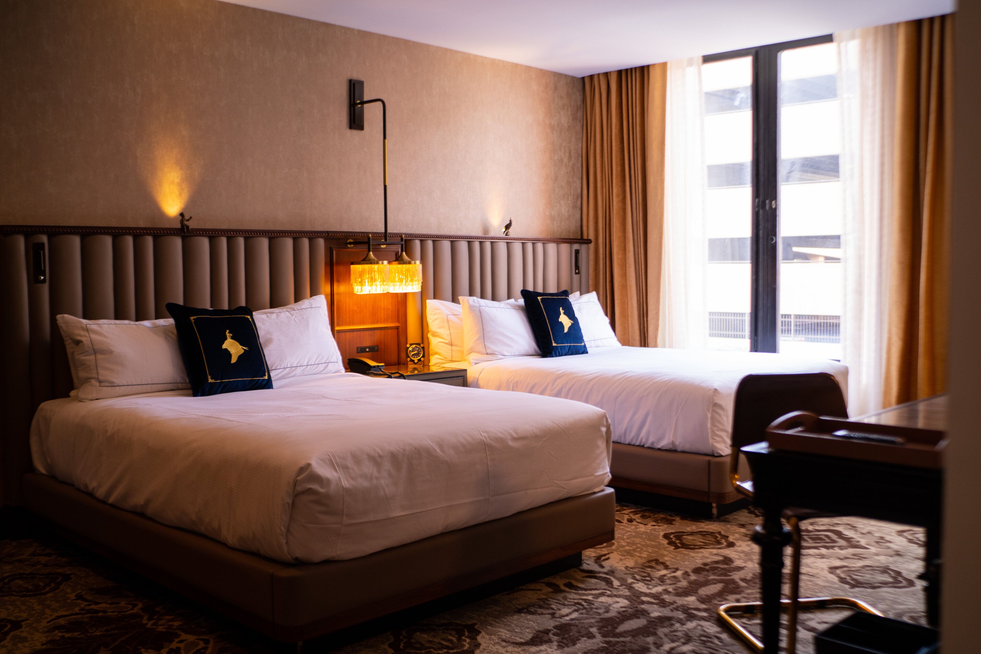 A hotel room with two beds, a desk, and a large window with curtains.