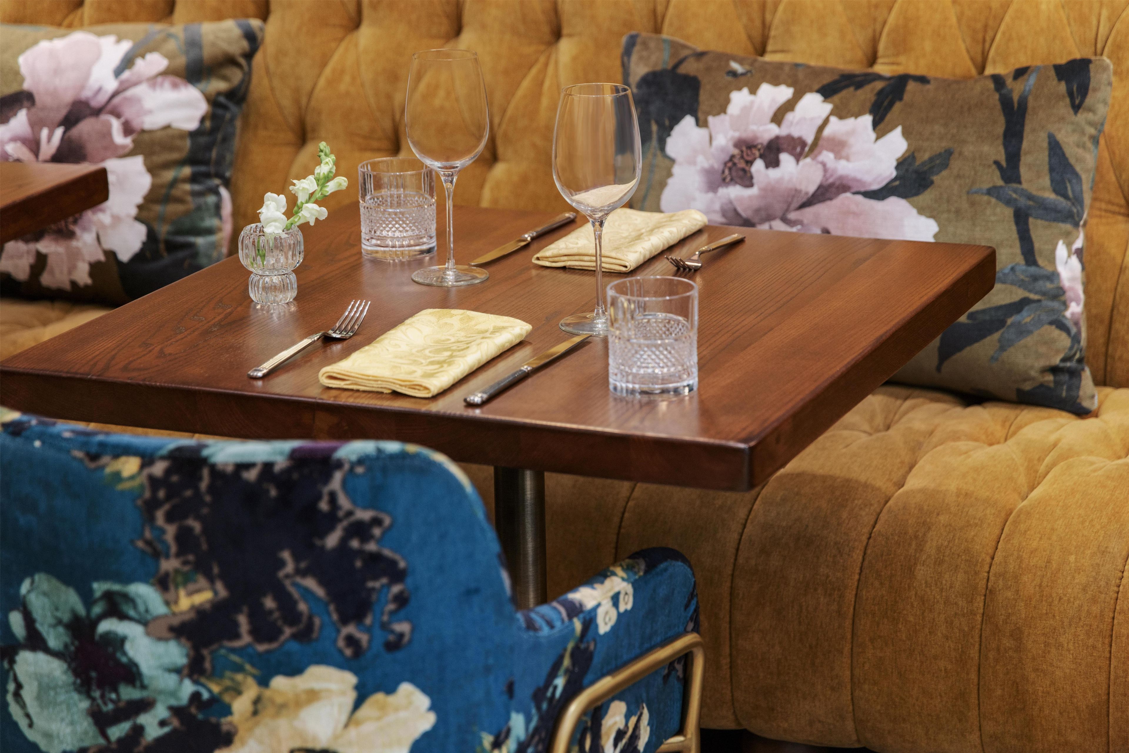 A dining table set with glassware, cutlery, and a napkin, placed in a room with a floral sofa and patterned cushions.