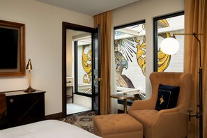 A hotel room with a bed, a desk, a chair, a lamp, and a view of a mural through the window. - The Peregrine Omaha Downtown, Curio Collection by Hilton (Omaha)