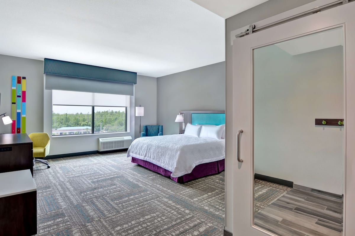 A modern hotel room with a large bed, a desk, a chair, and a view of the outdoors.