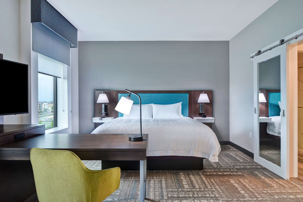 A hotel room with a bed, desk, chair, TV, lamps, and a large mirror.