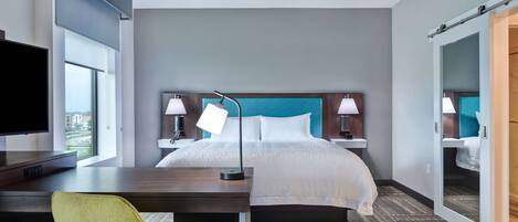 Habitación de hotel con cama, escritorio, silla, televisor, lámparas y un espejo grande.