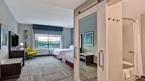 A hotel room with a bed, a desk, a chair, a shower, and a window with a view of greenery. - Hampton Inn & Suites Tampa Riverview Brandon (Tampa)
