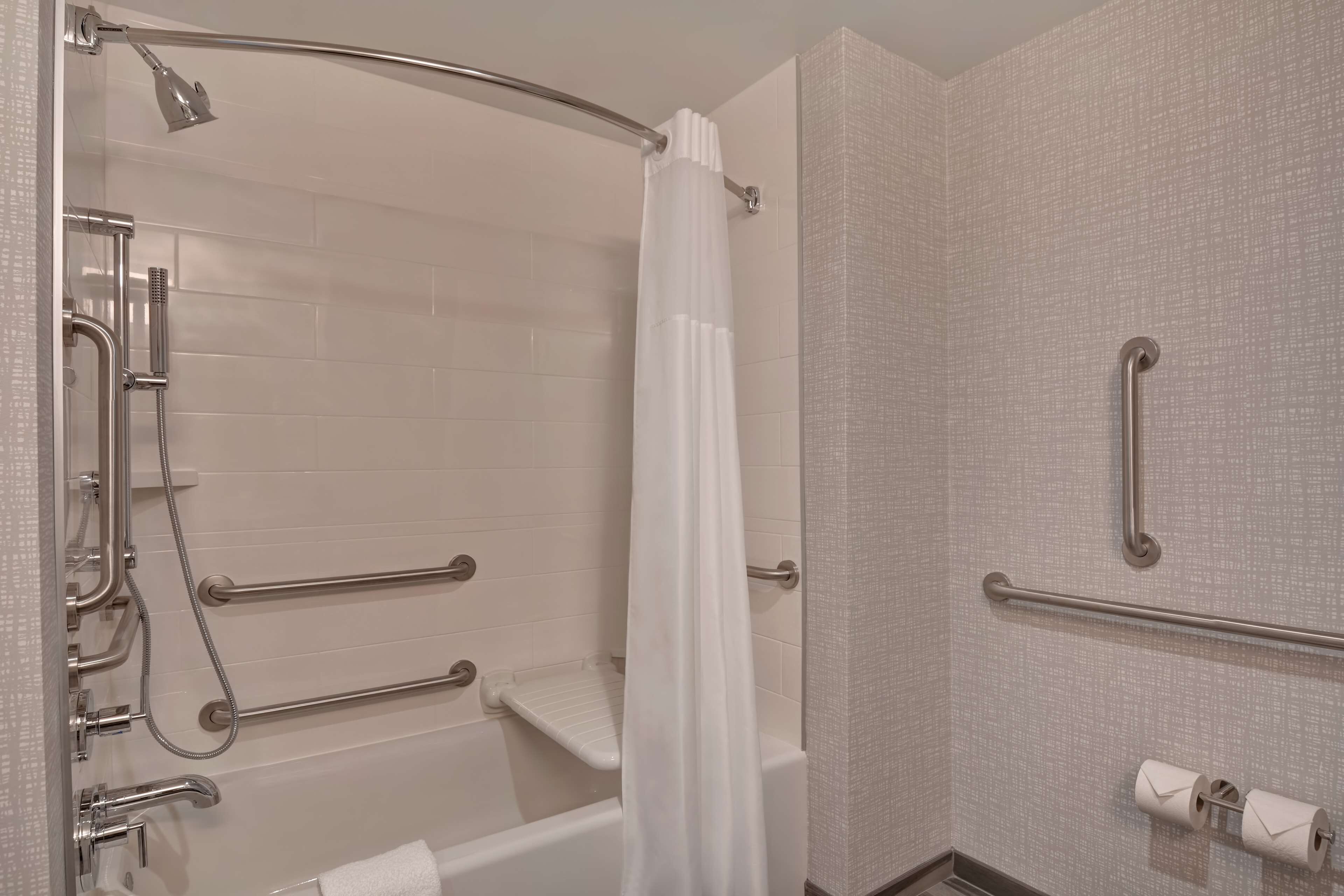 Une salle de bain moderne dotée d’une douche à l’italienne, de barres d’appui, d’un rideau de douche blanc et d’un porte-rouleau de papier toilette.