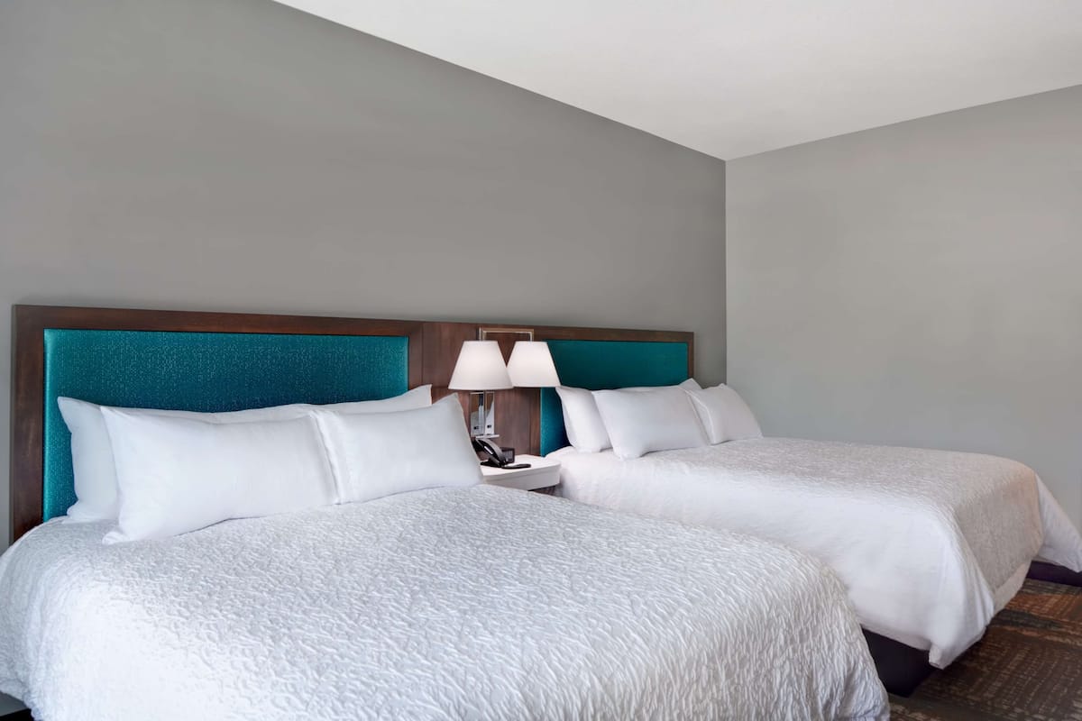 A hotel room with two beds, a teal headboard, white bedding, a bedside table with a lamp, and a phone.
