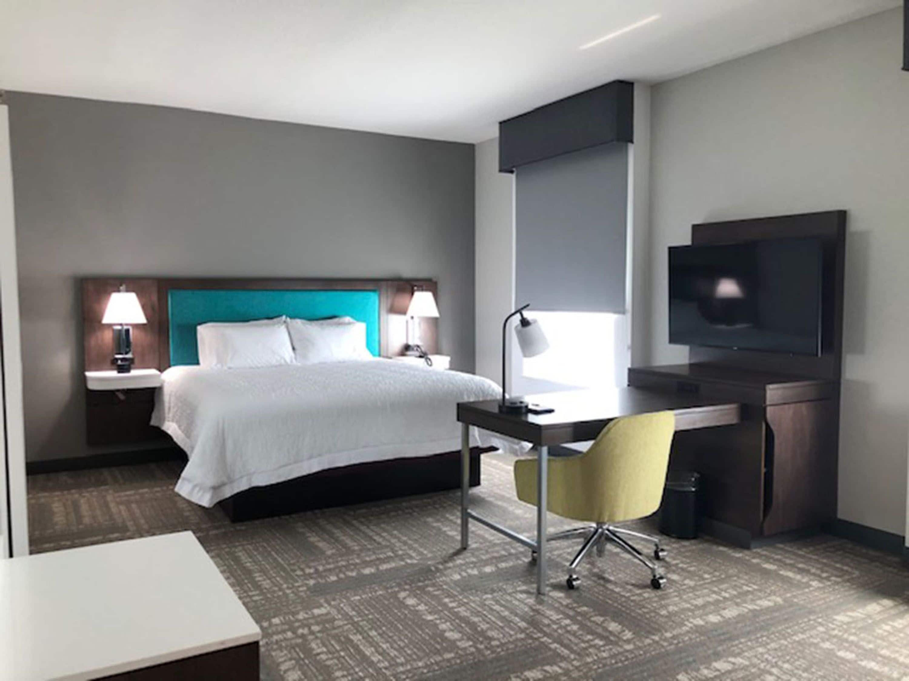 A hotel room with a bed, bedside tables with lamps, a desk with a chair, a flat-screen TV, and a large window.