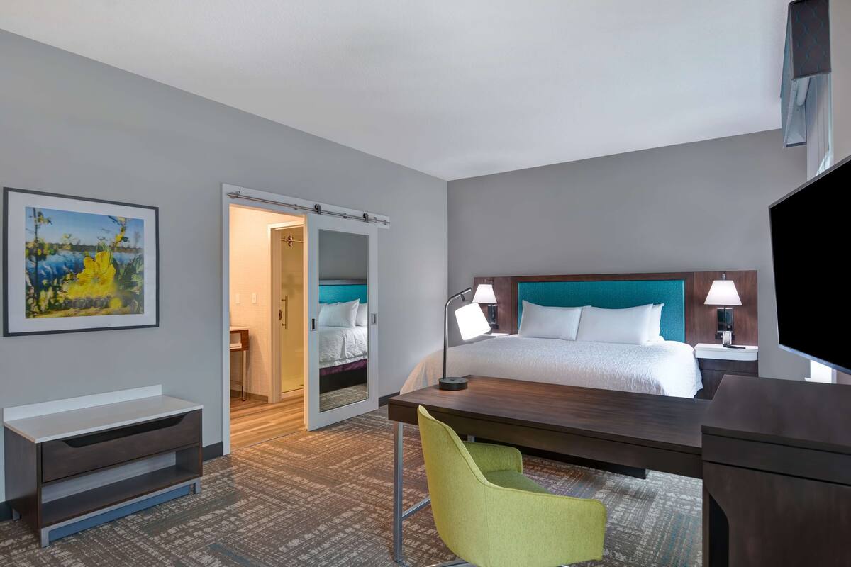 A hotel room with a bed, a desk, a chair, a TV, and a painting on the wall.