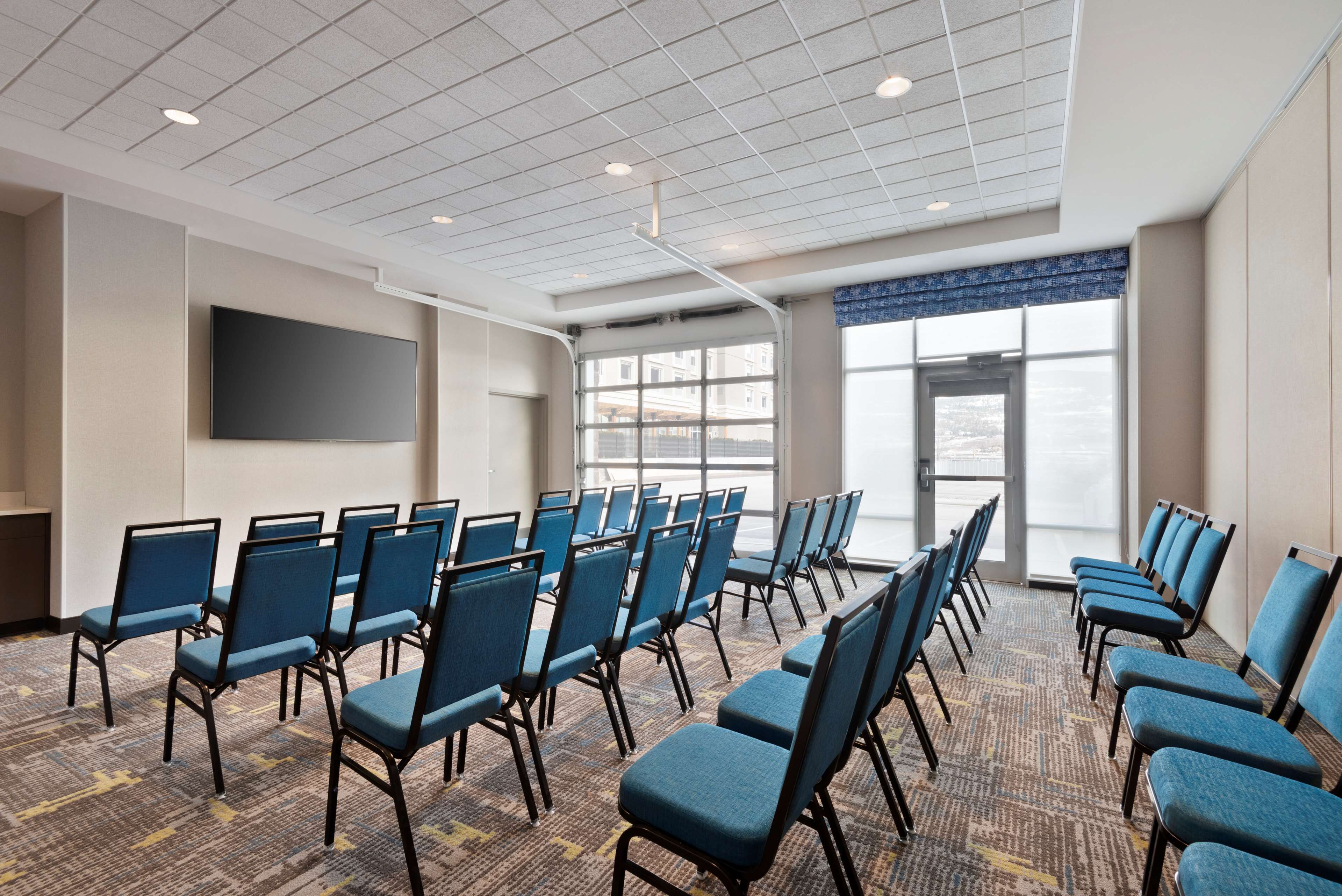 A conference room with rows of blue chairs, a large flat-screen TV on the wall, and large windows with a view of the city.