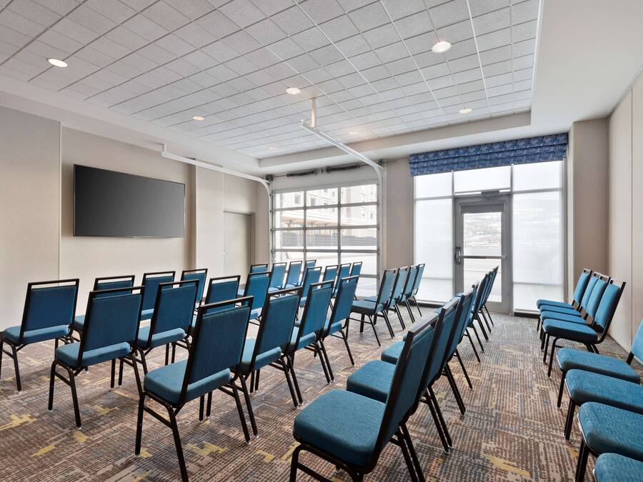 A conference room with rows of blue chairs, a large flat-screen TV on the wall, and large windows with a view of the city.