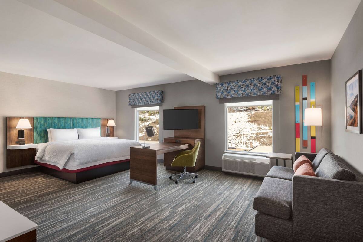 A hotel room with a bed, desk, chair, couch, and large windows.