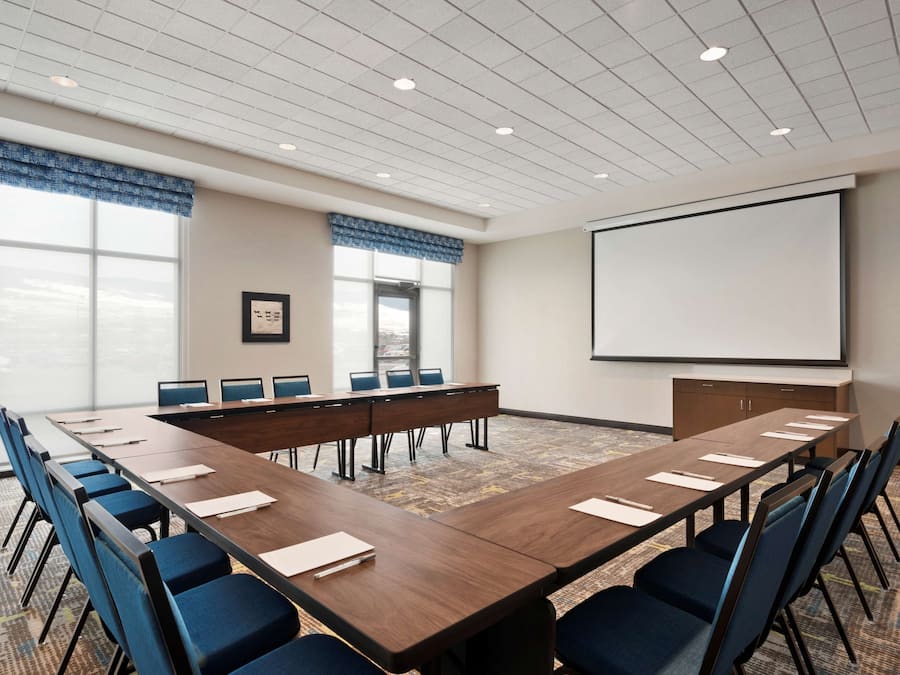 A conference room with a long table, blue chairs, a projector screen, and large windows with blue valances.
