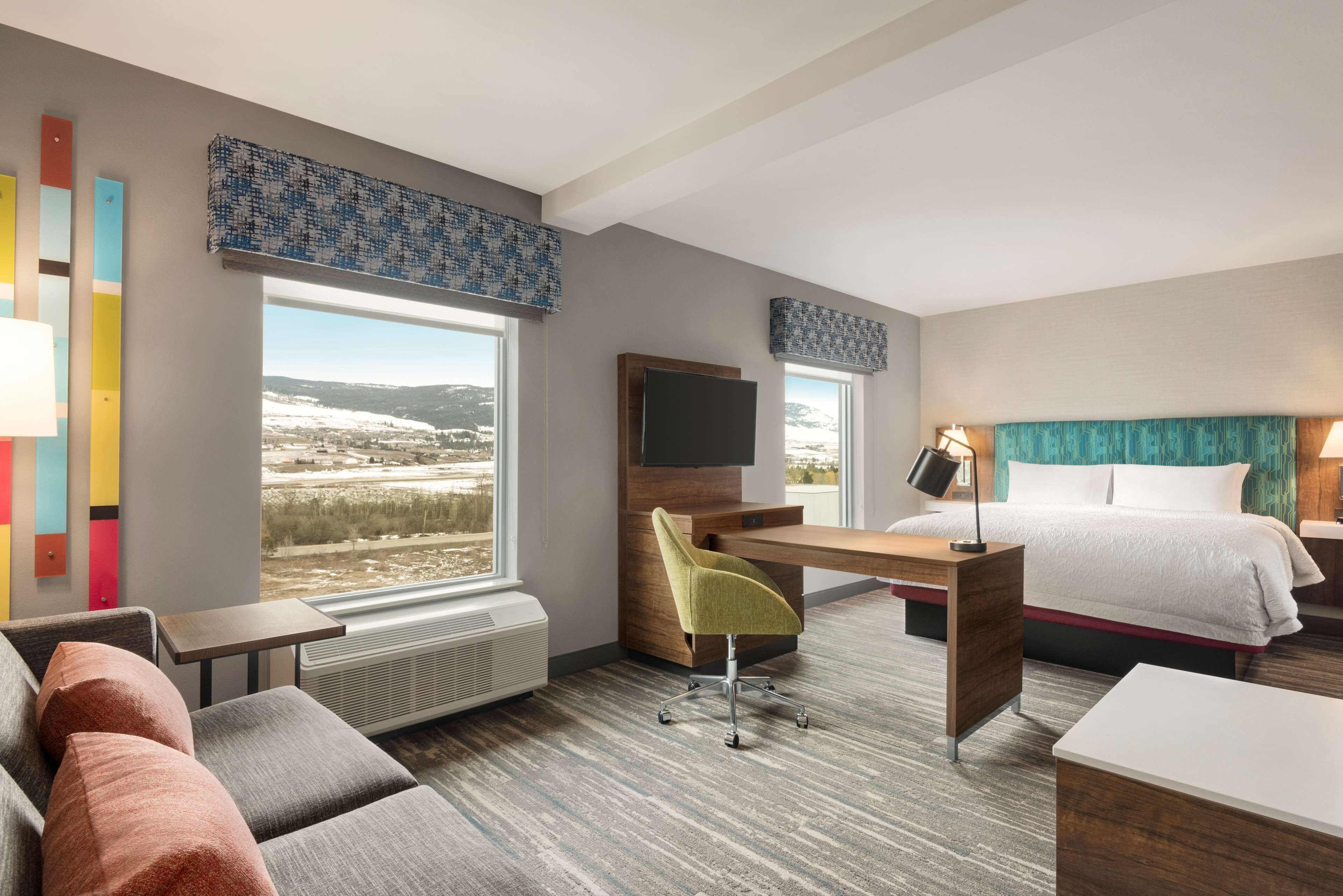 A modern hotel room with a desk, a comfortable sofa, a bed, and large windows offering a view of the outdoors.